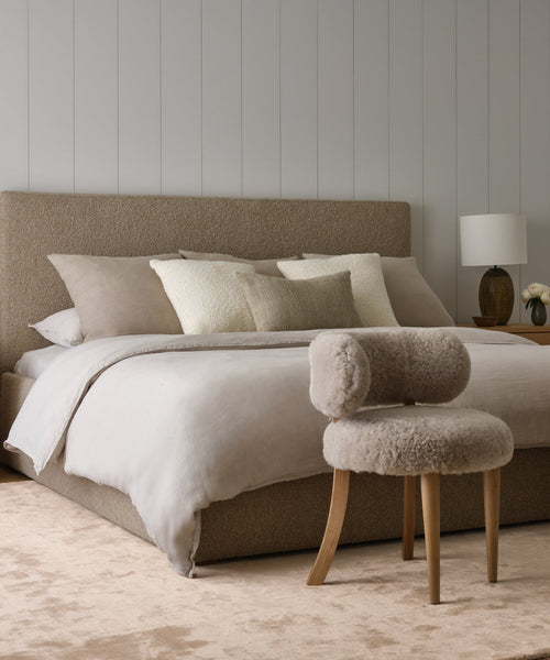 A modern bedroom features a beige upholstered bed, neutral bedding, a fuzzy taupe chair, wooden nightstand with lamp and flowers, all atop the Bristol Rug—handwoven in India—layered over light carpet.