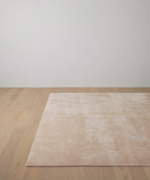 The Bristol Rug, a large handwoven piece in light pink with a soft texture, is placed on a light wooden floor in a minimalistic room with white walls.