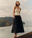 Woman wearing a cream top and black skirt standing on a rooftop with a scenic view of mountains and sea.