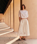 Woman in a white outfit standing in front of a beige wall with sunlight casting shadows.