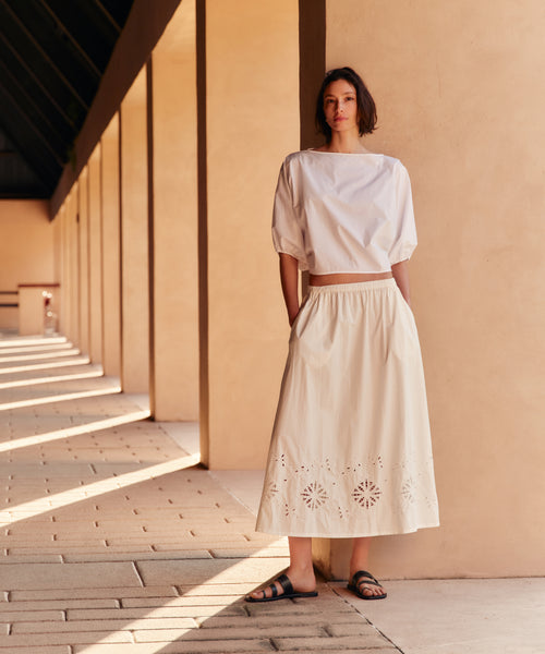 Woman in a white outfit standing in front of a beige wall with sunlight casting shadows.