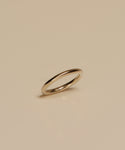The Brook Gold Band, a sleek and thin 14k gold ring, stands upright on a smooth beige surface, casting a subtle shadow below it.
