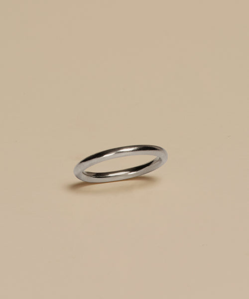 The Brook Gold Band, a slim white gold ring, rests on a plain beige surface with a subtle shadow beneath.
