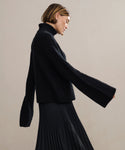 A woman with short, light brown hair wears the black Cable Nell Turtleneck and a black pleated skirt, standing in profile against a neutral background.