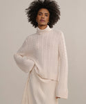 A woman with curly hair stands against a neutral background, wearing the Cable Nell Turtleneck sweater with a boxy silhouette and an ivory pleated skirt.