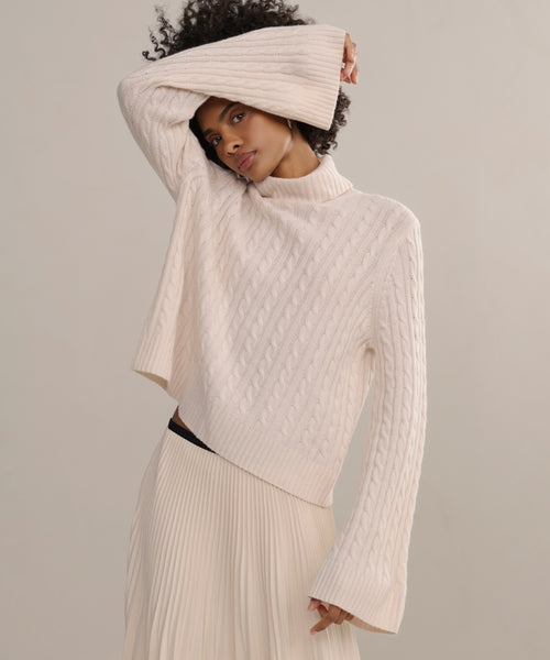 A woman models the Cable Nell Turtleneck, an ivory cable-knit sweater with flared sleeves, paired with a pleated skirt. She poses with one arm raised, partially covering her face against a plain light background.