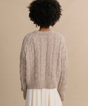 A person with short, curly hair is seen from behind wearing the Cable Serena Crewneck—a chunky beige alpaca-wool sweater—with a cream skirt, all set against a plain beige background.