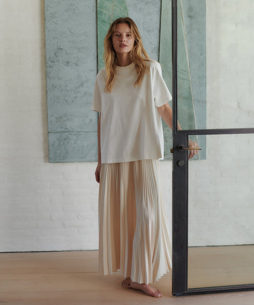 A woman stands indoors wearing an oversized cream short-sleeve top and a long pleated cream skirt, holding the door frame of a glass door. The background features a white brick wall and light wood flooring.
