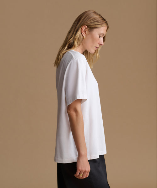 A woman in profile wearing a loose white short-sleeve t-shirt and black pants, standing against a solid beige studio background.