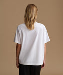 A woman viewed from the back wearing an oversized white short-sleeve T-shirt and black pants, standing against a plain warm beige studio background.