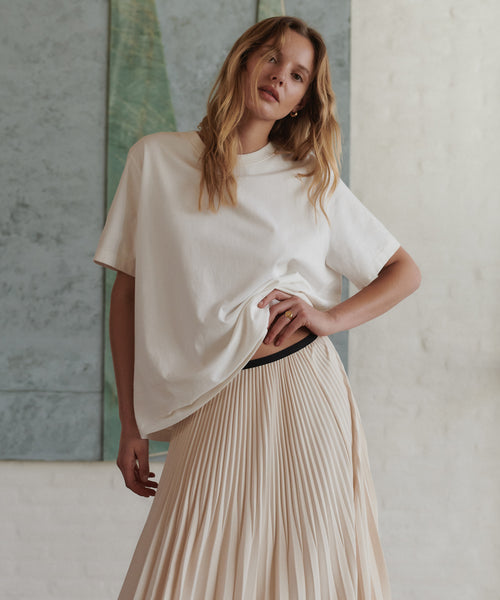 A woman modeling an oversized off-white short-sleeve top and a pleated light beige skirt, standing against a textured light gray wall.
