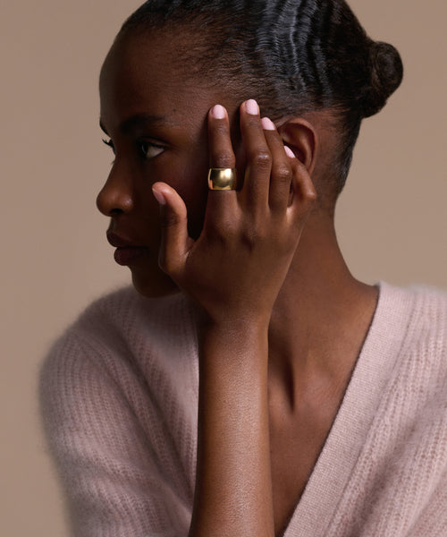 A woman in a soft pink sweater poses in profile against a neutral background, resting her hand on her face and wearing the Campbell Cigar Band. Her hair is styled in a sleek bun.