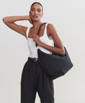 A woman in a white tank top and high-waisted black pants poses with one hand behind her neck, carrying the stylish Canvas Day Bag over her shoulder against a plain light background.