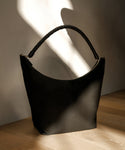 A Canvas Day Bag in classic black with a curved top and thick, twisted handle rests on a wooden floor, bathed in soft natural light and shadow.