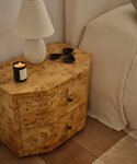 A cozy bedside scene features the Carbon Nightstand in light wood with natural grain, a lit candle, white lamp, and small black dishes. Cream bedding adds warmth beside the nightstand’s surface and drawers.