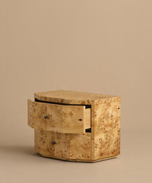 The Carbon Nightstand is a small, modern piece crafted from light, textured burlwood with a natural pattern. It features two drawers, one partially open, set against a plain beige background.