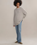 A person with curly hair stands against a light background, wearing an oversized light gray Cashmere Amelia Crewneck, blue jeans, and brown shoes. One hand is behind their back as they gaze slightly to the side.