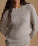 A close-up of a person wearing a light grey knit crewneck sweater against a solid warm beige background.