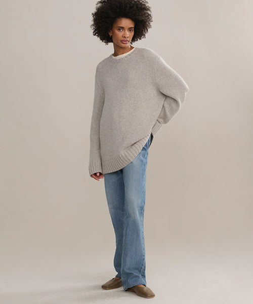 A person with curly hair stands against a light background, wearing an oversized light gray Cashmere Amelia Crewneck, blue jeans, and brown shoes. One hand is behind their back as they gaze slightly to the side.
