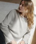 A woman wearing a light gray knit crewneck sweater, shown in a close-up lifestyle shot with warm natural light.