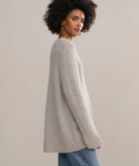 A woman with curly hair stands sideways against a plain light background, wearing the oversized Cashmere Amelia Crewneck in light gray and blue jeans while looking over her shoulder.
