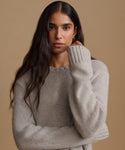 A woman with long dark hair wearing a light grey knit crewneck sweater, posing against a solid warm beige background.