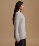 A woman in side profile wearing a light grey knit sweater and a black pleated skirt, set against a solid tan studio background.