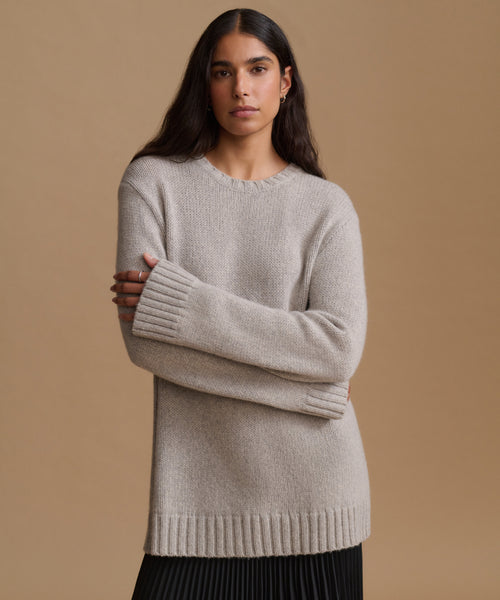 A woman modeling a light beige knit crewneck sweater with ribbed cuffs and hem, paired with a black pleated skirt, against a solid tan background.