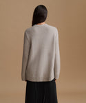 A woman shown from the back wearing a light grey knit sweater and a black pleated skirt, set against a solid warm tan studio background.