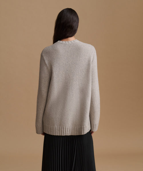 A woman shown from the back wearing a light grey knit sweater and a black pleated skirt, set against a solid warm tan studio background.