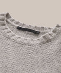 A close-up of the Cashmere Amelia Crewneck, a grey cashmere knit sweater with a ribbed crew neckline and black label inside the collar, displayed on a neutral beige background.