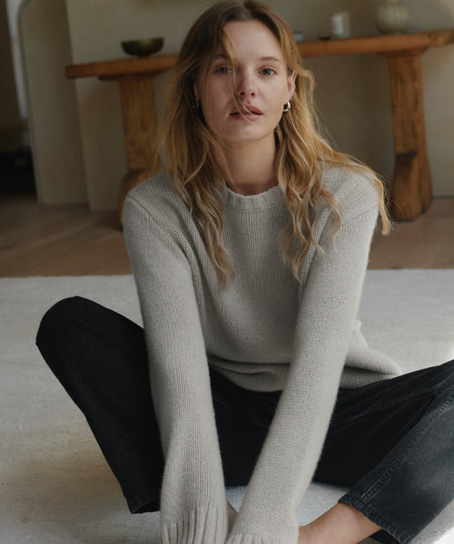 A woman with blonde hair sits cross-legged on a white rug in a warm room, wearing a light grey ribbed knit sweater and black jeans.