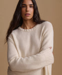 A woman wearing a cream-colored, textured knit crewneck sweater, posed against a solid warm beige background.