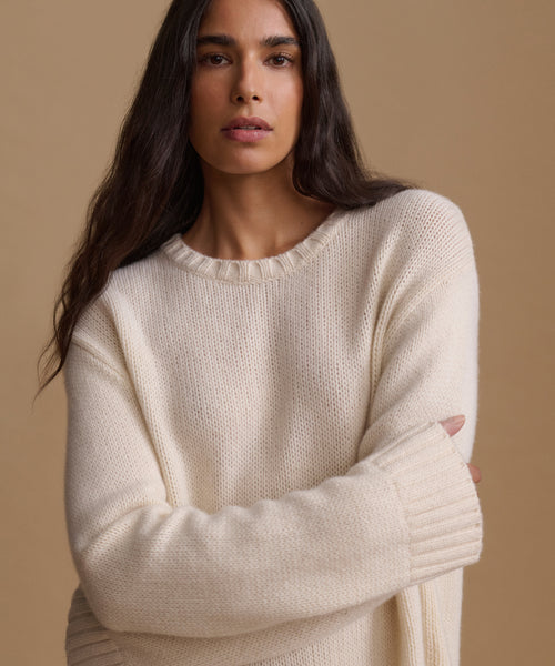 A woman wearing a cream-colored, textured knit crewneck sweater, posed against a solid warm beige background.