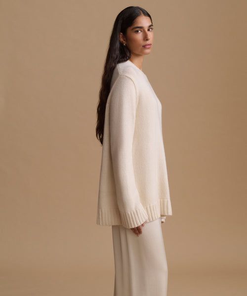 A woman stands in profile wearing a cream knit sweater and matching knit pants against a plain beige studio background.
