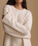 A close-up of a woman wearing a cream-colored knit sweater with a crew neck and ribbed cuffs, posed against a solid beige background.