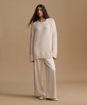 A woman stands against a warm beige studio background wearing an oversized cream knit sweater and matching wide-leg knit pants.