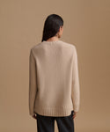 A woman viewed from the back wearing a light beige knit sweater paired with black pants, against a solid tan studio background.