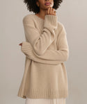 A person with curly hair wears the Cashmere Amelia Crewneck, a loose, lightweight beige sweater with long sleeves, standing against a neutral background and resting one hand on their chin.