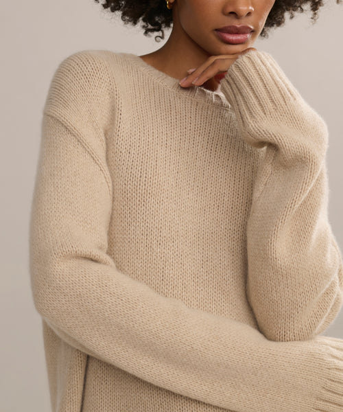 Wearing the Cashmere Amelia Crewneck, a person stands with one hand near their chin, highlighting the soft texture and long, loose sleeves of the beige oversized sweater. The image captures only their upper body.