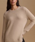 A close-up of a woman wearing a beige ribbed knit crewneck sweater against a solid warm beige background.