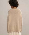 A person with short curly hair is shown from behind, wearing the Cashmere Amelia Crewneck in light beige and a light pleated skirt, set against a plain, neutral background.