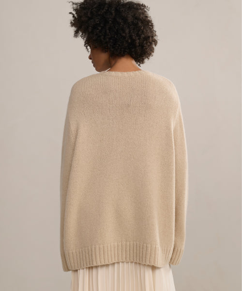 A person with short curly hair is shown from behind, wearing the Cashmere Amelia Crewneck in light beige and a light pleated skirt, set against a plain, neutral background.