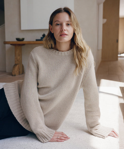 A woman wearing a cream-colored ribbed knit sweater, kneeling on a light-colored rug in a minimalist indoor setting.
