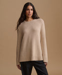 A woman modeling a beige crewneck knit sweater paired with black pants, against a solid beige studio background.