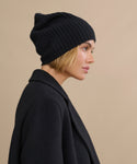 A woman with a short blonde bob wears the Cashmere Beanie in black and a matching coat, shown in profile against a beige background.