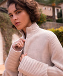 A woman with short, wavy brown hair wears the Boyd Cashmere Sweater, a beige fisherman rib zip-up style, partly unzipped to reveal a silver ring. She stands outside in sunlight with plants and buildings behind her, looking at the camera.