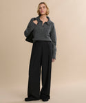 A woman with short blonde hair and a neutral expression stands against a beige background wearing the Cashmere Burke Polo, loose black trousers, black shoes, and carrying a black bag over her shoulder.
