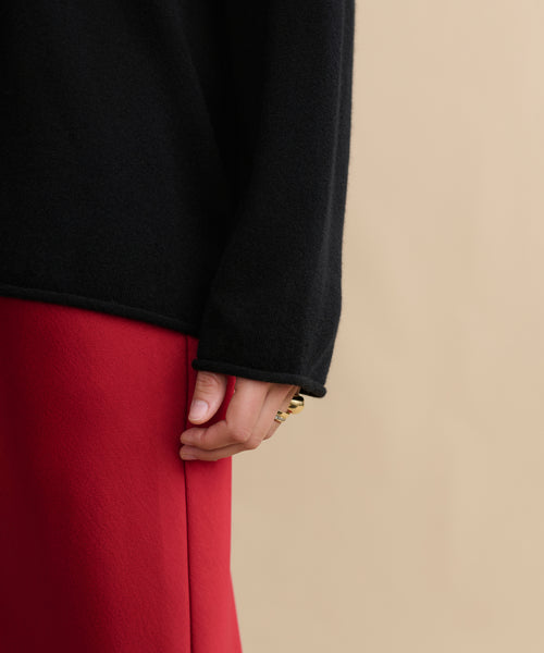 Wearing the black Cashmere Chandler Long-Sleeve Tee and a red skirt, a person stands against a beige background with their hand near their hip, showcasing gold rings.