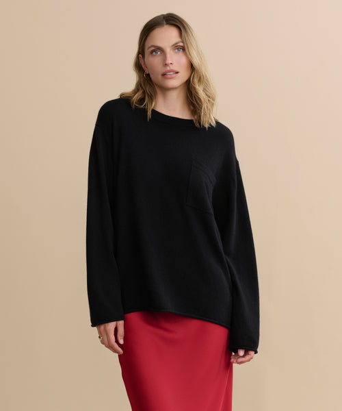 A woman with wavy blonde hair wears the Cashmere Chandler Long-Sleeve Tee with a chest pocket and a red skirt, standing against a plain beige background.